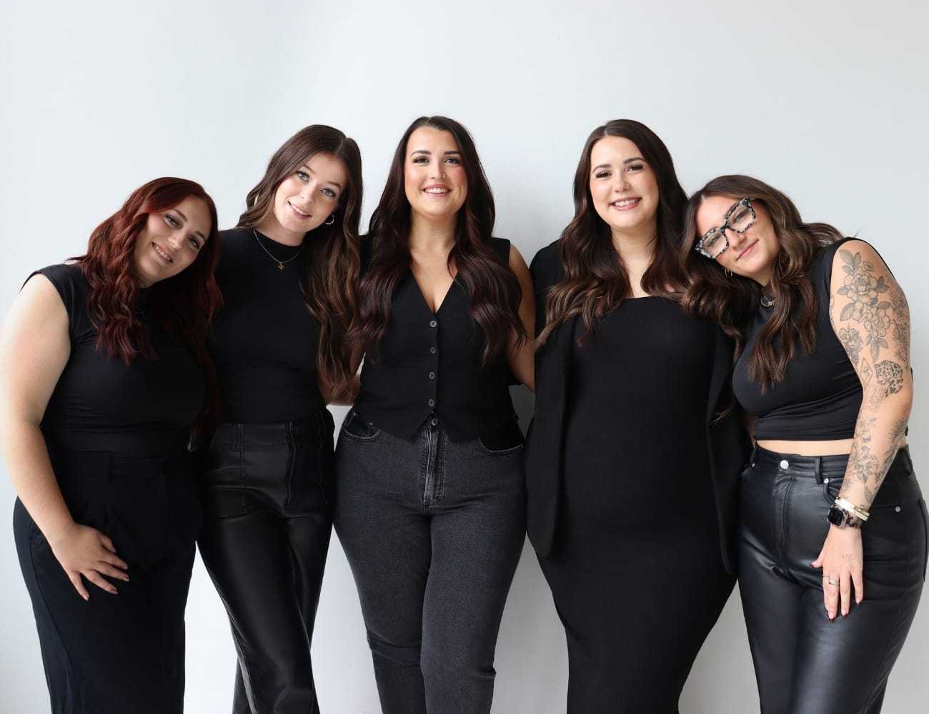 Five women in black outfits smiling and posing against a plain white background.